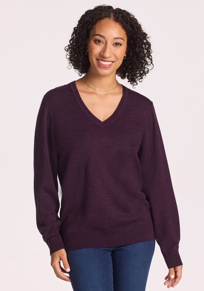 A woman with curly hair smiles in blue jeans and the Woolx Francesca Sweater in Deep Plum, standing against a plain, light background. 