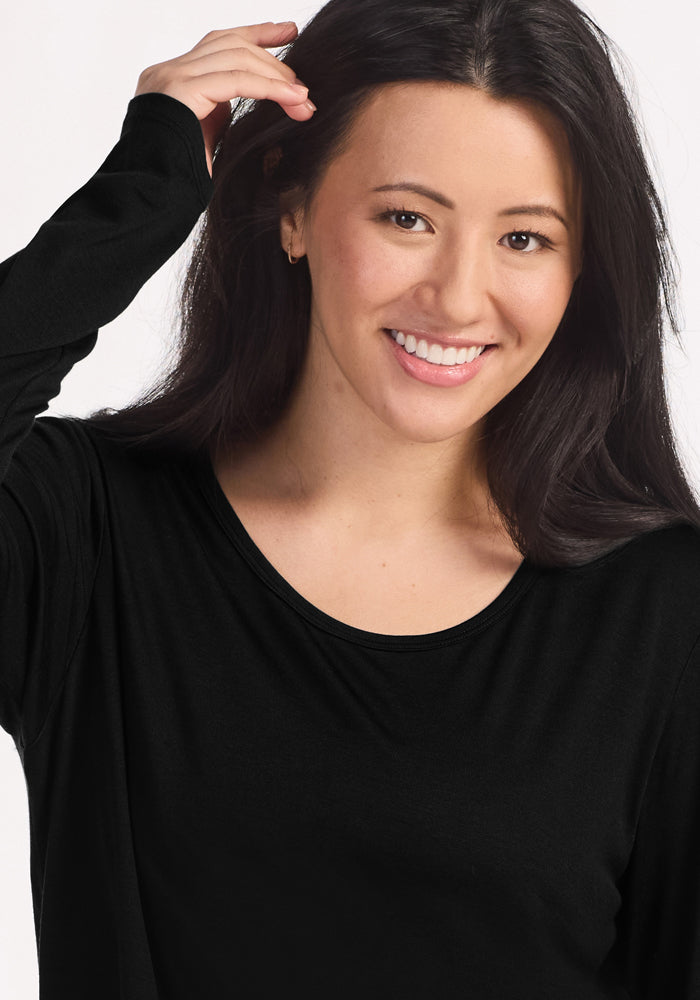 A woman with long dark hair smiles warmly at the camera, modeling the Woolx Lucille Long Sleeve Nightgown in black, with one hand raised near her head against a plain light background.