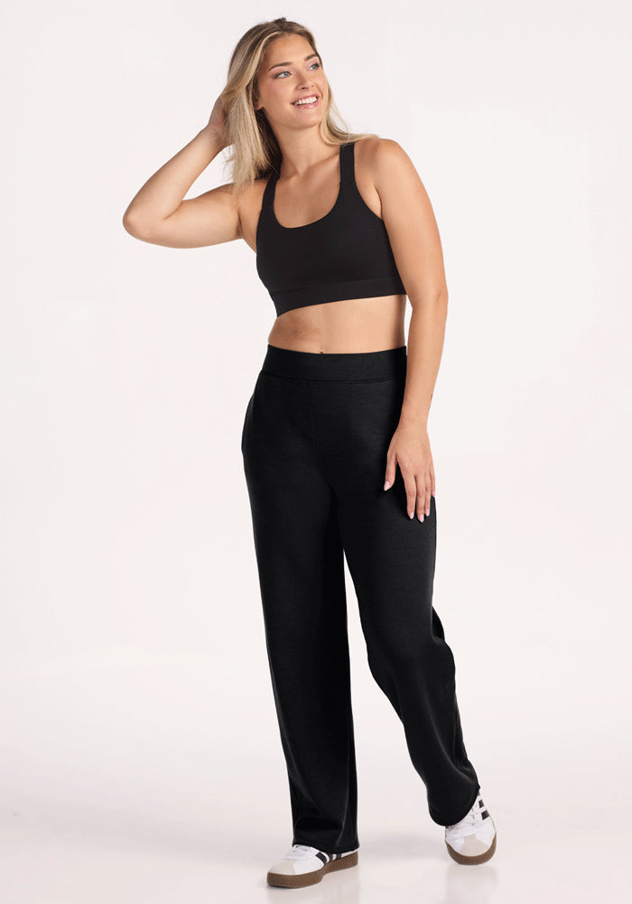 A woman with long blonde hair smiles and touches her hair while wearing Woolx Bree Lounge Pants in Black, a black sports bra, and white sneakers. She stands against a plain white background, looking to the side.