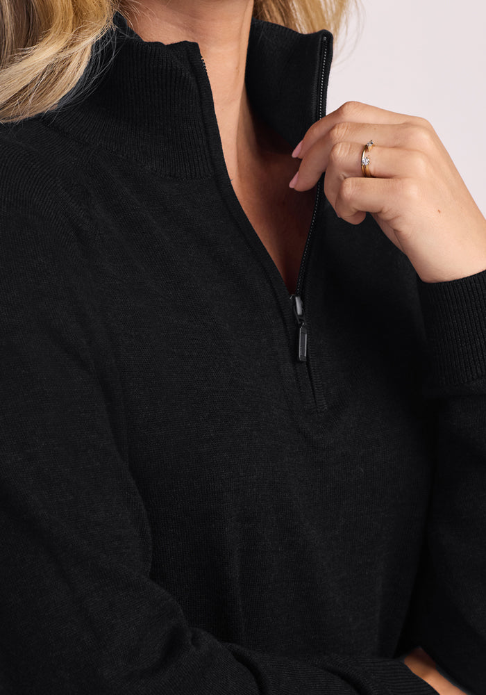 Wearing the Woolx Collins Half Zip Sweater in black, a person touches their chin, revealing a gold ring with a small gemstone on their ring finger; only the lower face and upper body are visible.