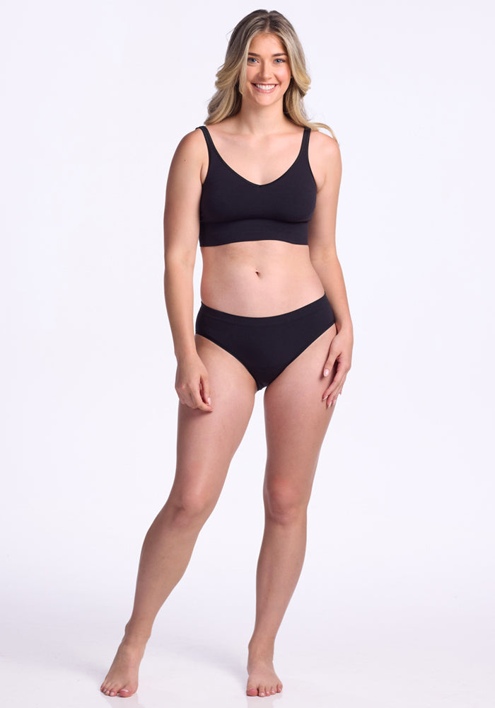 A woman with long, wavy blonde hair smiles barefoot against a white background, wearing the Woolx Maeve Bikini - Black, a seamless set made from moisture-wicking fabric.