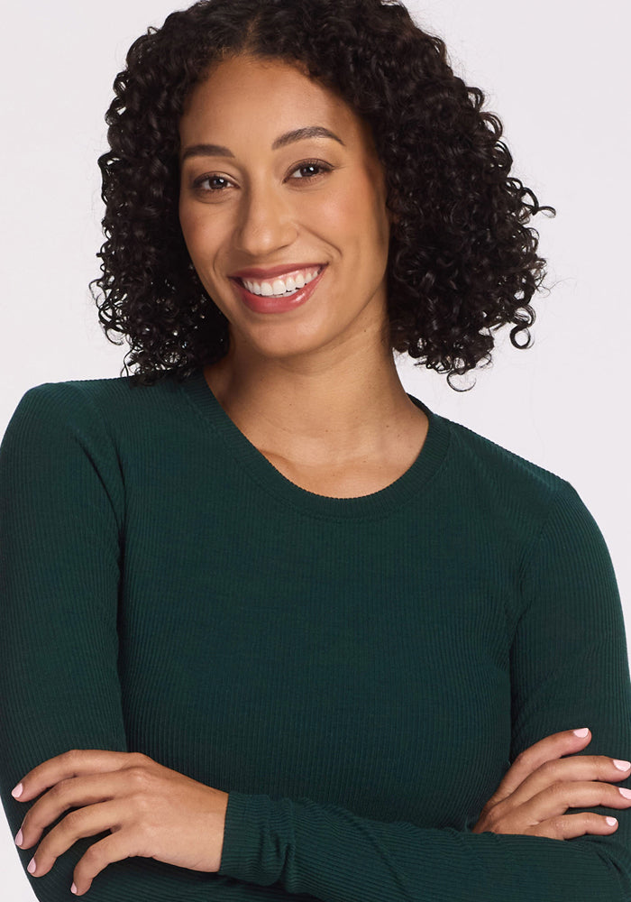 A woman with curly dark hair smiles with her arms crossed, wearing the Woolx Hadley Ribbed Crew in Deep Timber against a plain light background.