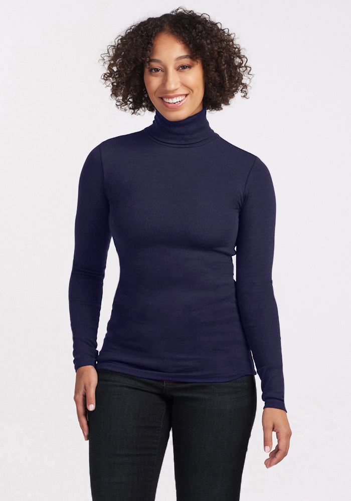 A woman with curly hair smiles while wearing the Woolx Sage Ribbed Turtleneck in Midnight Sapphire and dark jeans, standing against a plain white background. 