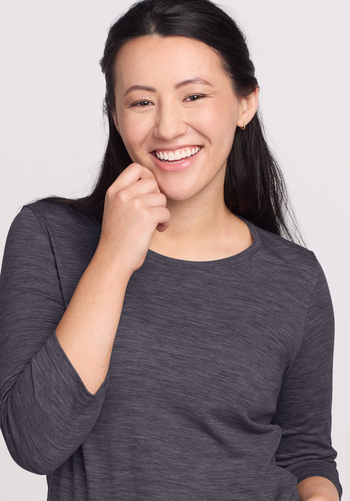 A woman with long dark hair smiles, wearing the Woolx Jenny 3/4 Sleeve Crew Neck in Cinder Melange, her hand near her chin against a light background.