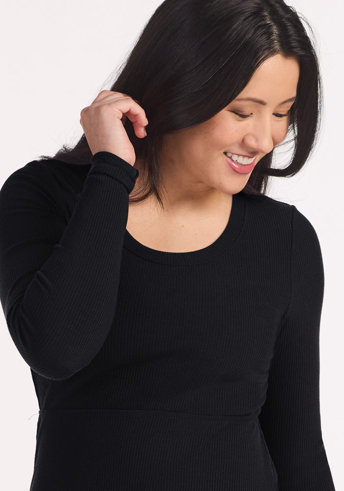 Woman with long dark hair smiles, wearing the Woolx Caroline Ribbed Twirl Dress in black and touching her hair.