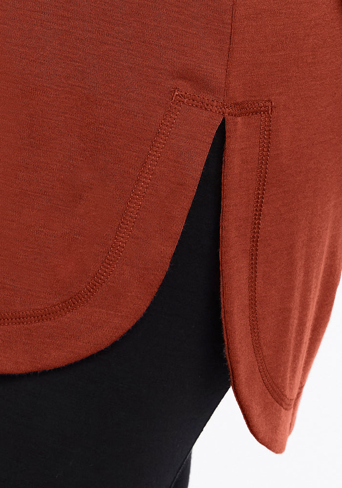 Close-up of the Woolx Blair Tunic in Maple Leaf, featuring a rust hue, curved hem, and side slit. Merino wool fabric and detailed stitching are visible; styled over black pants.
