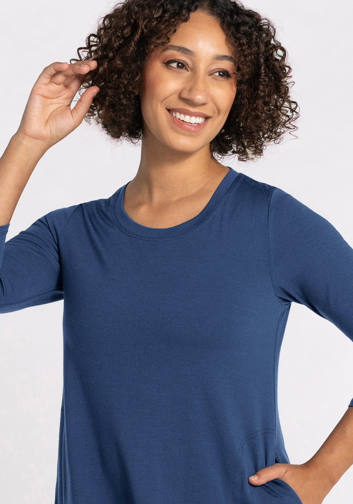 A woman with curly hair smiles, looking off to the side. She wears the Woolx Rissa A Line Dress in Starry Night—its long-sleeved blue merino wool design is ideal for layering in fall and winter. One hand is raised by her head, the other at her waist. The background is plain and light.