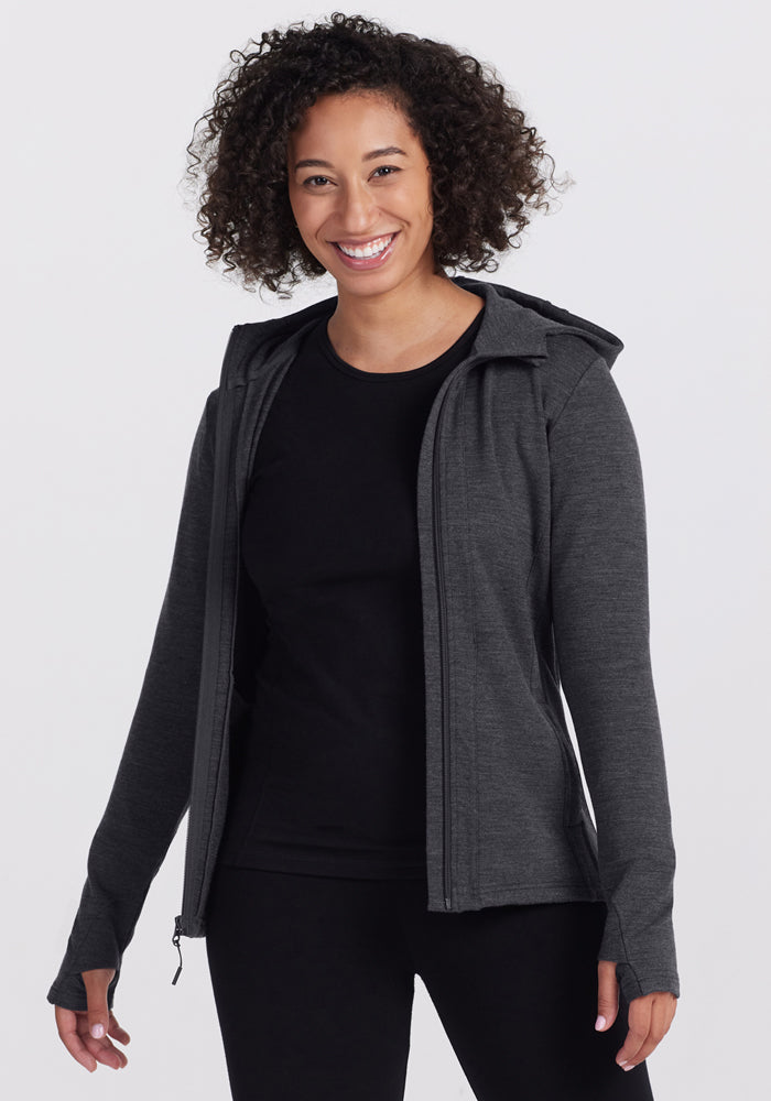 A curly-haired woman smiles wearing a Woolx Zoey Zip Up Hoodie in Cinder Melange over a black shirt and black pants, standing against a plain light background. | Tori is 5'7", wearing a size S