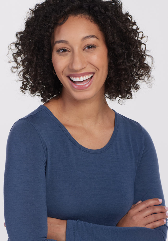 A woman with curly hair smiles warmly, arms crossed, wearing the Woolx Remi Long Sleeve T Shirt in Starry Night against a plain light background.
