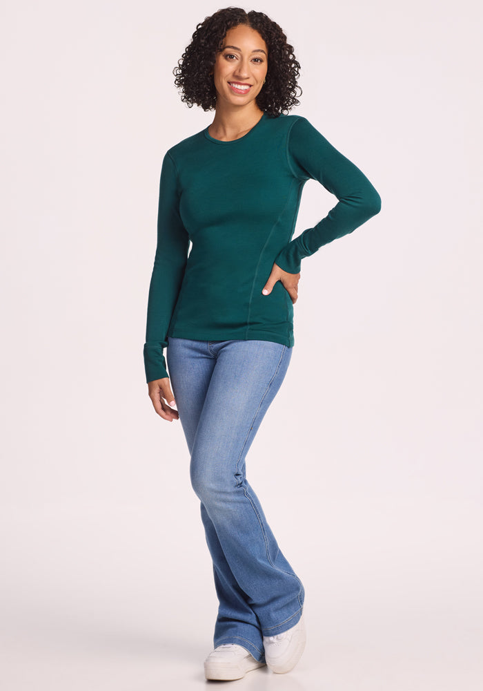 A woman with curly hair smiles while wearing the Woolx Hannah Long Sleeve Top in Riverbank Green, paired with blue jeans and white shoes, standing against a plain light background.