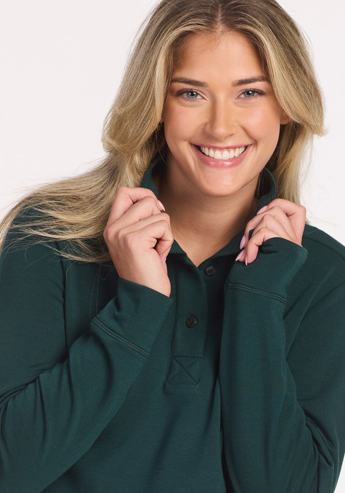 A smiling woman with long blonde hair wears the Woolx Annie 3 Button Pullover in Deep Timber, holding the collar with both hands as she looks directly at the camera against a plain white background.