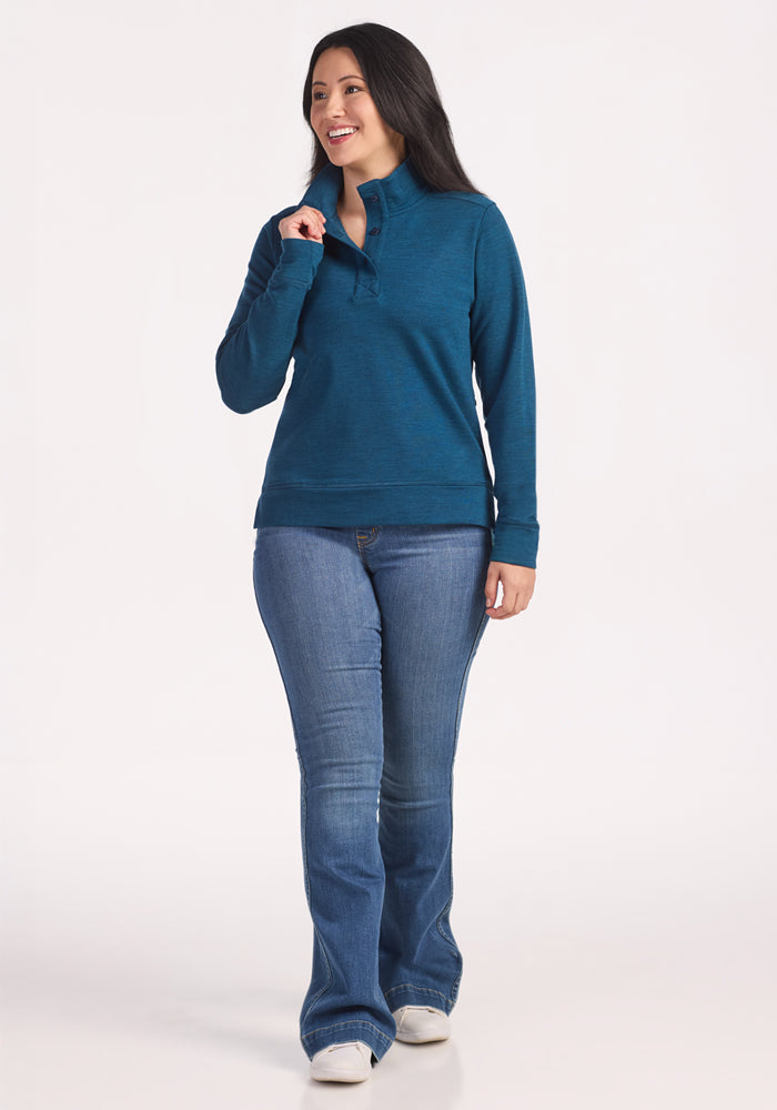 A woman with long dark hair smiles against a white background, wearing the Woolx Annie 3 Button Pullover in Majolica Blue and blue jeans. She stands with her right hand near her collar, looking slightly to her left.