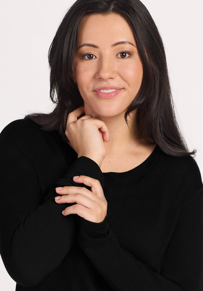 A woman with long dark hair, wearing the Woolx Ashlie Lounge Top in black, smiles gently with one hand on her neck and the other on her wrist against a plain, light background.