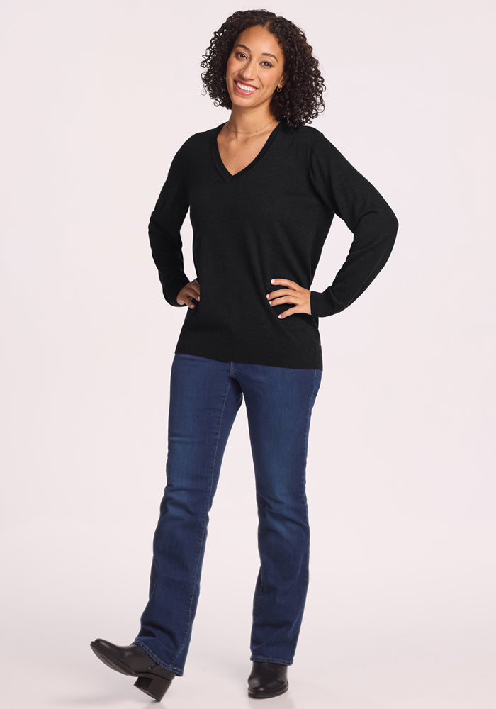 A smiling woman with curly hair wears the Woolx Francesca Sweater in black, blue jeans, and black shoes. She stands with one hand on her hip and one foot slightly raised against a plain light background.
