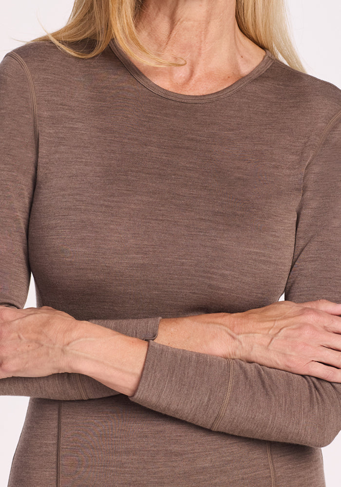 A woman with light skin and blonde hair is shown from the shoulders down, wearing the Woolx Hannah Long Sleeve Top in Simply Taupe. She stands with her arms crossed; her face is not visible.