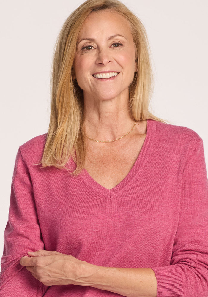 A smiling woman with long blonde hair wears the Woolx Francesca Sweater in Wild Salmon and a delicate gold necklace, standing against a plain light background.