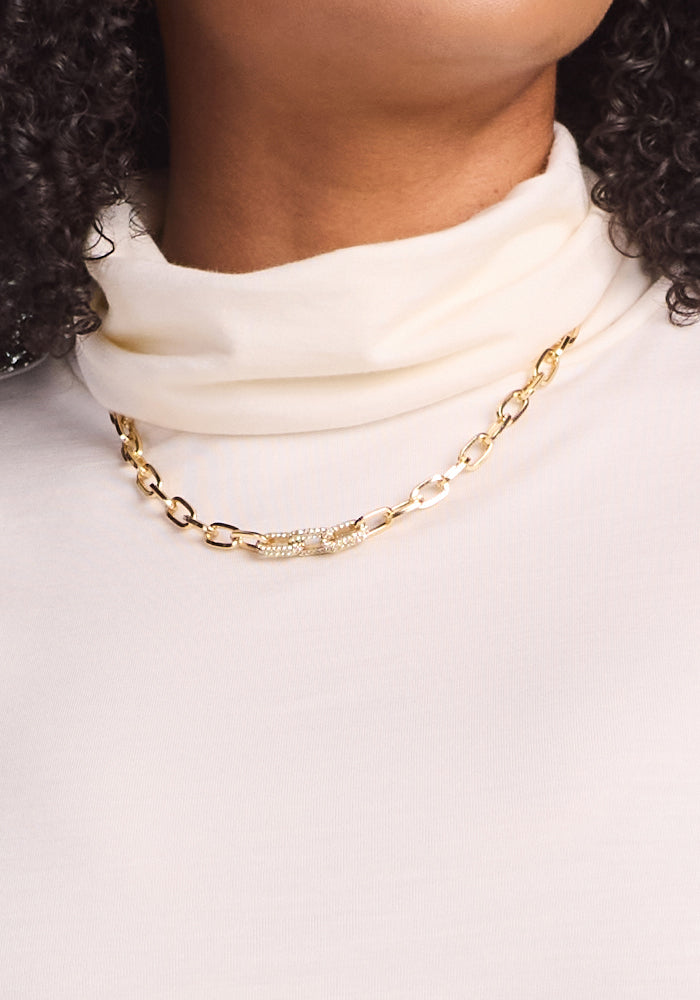 A close-up of a person with curly hair, partially visible, wearing the Woolx Kennedy Bodysuit in Buttercream and a gold chain necklace featuring a crystal-encrusted curved bar—an essential layering look.