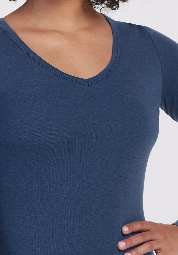 A person wearing the Woolx Layla V Neck Top in Starry Night, a fitted long-sleeve blue Merino wool shirt, stands with one hand on their hip against a plain background.