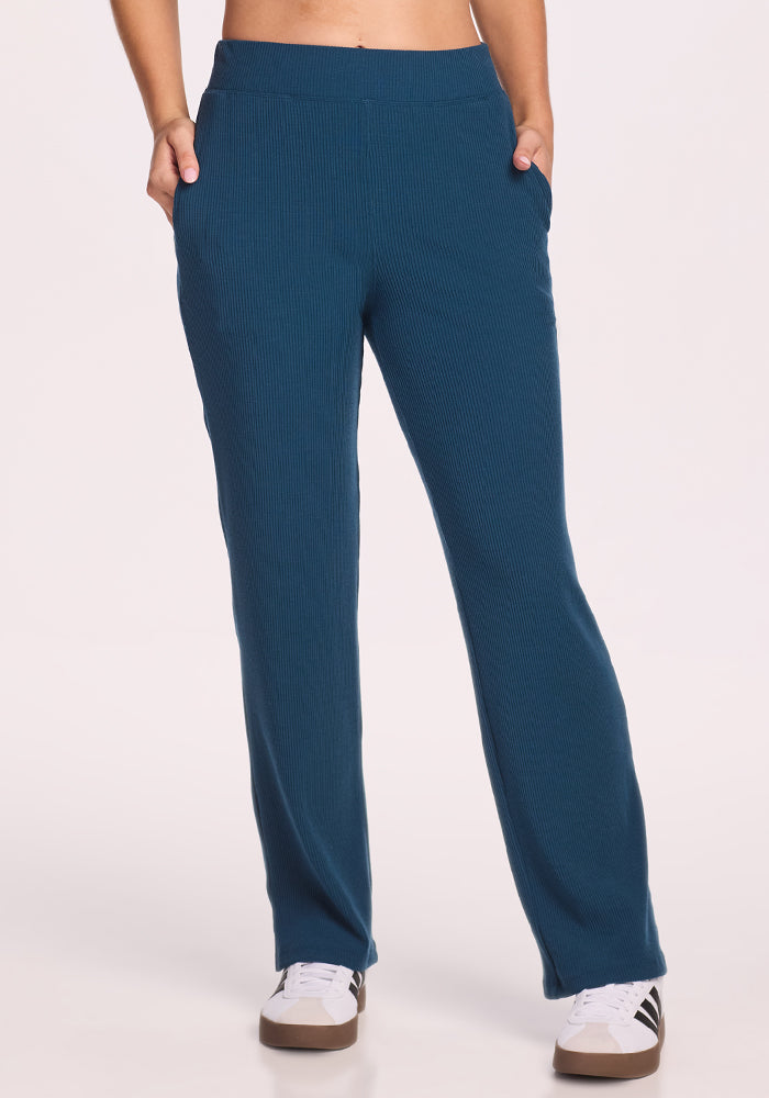 A person wears Woolx Luca Ribbed Pants in Majolica Blue with hands in pockets, paired with white sneakers with black stripes, standing against a plain light background. The wide-leg pants offer a relaxed, stylish look for lounging or casual outings. 