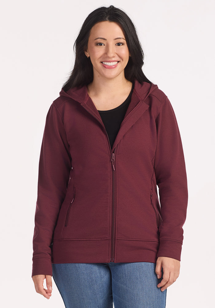 A woman with long dark hair smiles at the camera, wearing the Woolx Darby Zip Up Hoodie in Toasted Garnet over a black shirt and blue jeans, standing against a plain light background. 