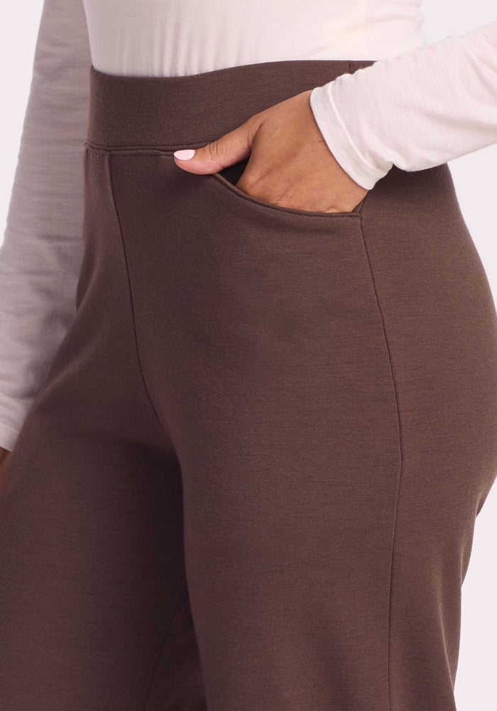 A petite woman models the Woolx Ellie Wide Leg Pants Petite in Bracken, styled with a white long-sleeve top and one hand tucked in her pocket—shown from waist down.