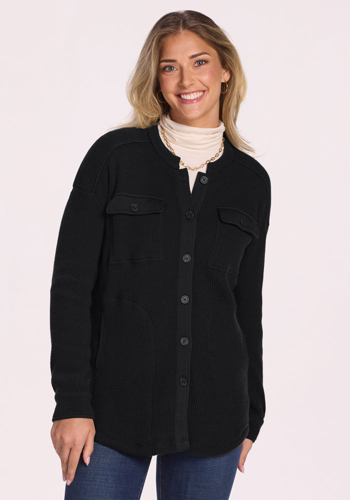 A woman with long, wavy blonde hair smiles in the Woolx Oaklynn Waffle Shacket in black, styled over a light turtleneck and blue jeans with a gold necklace, set against a plain light background. 