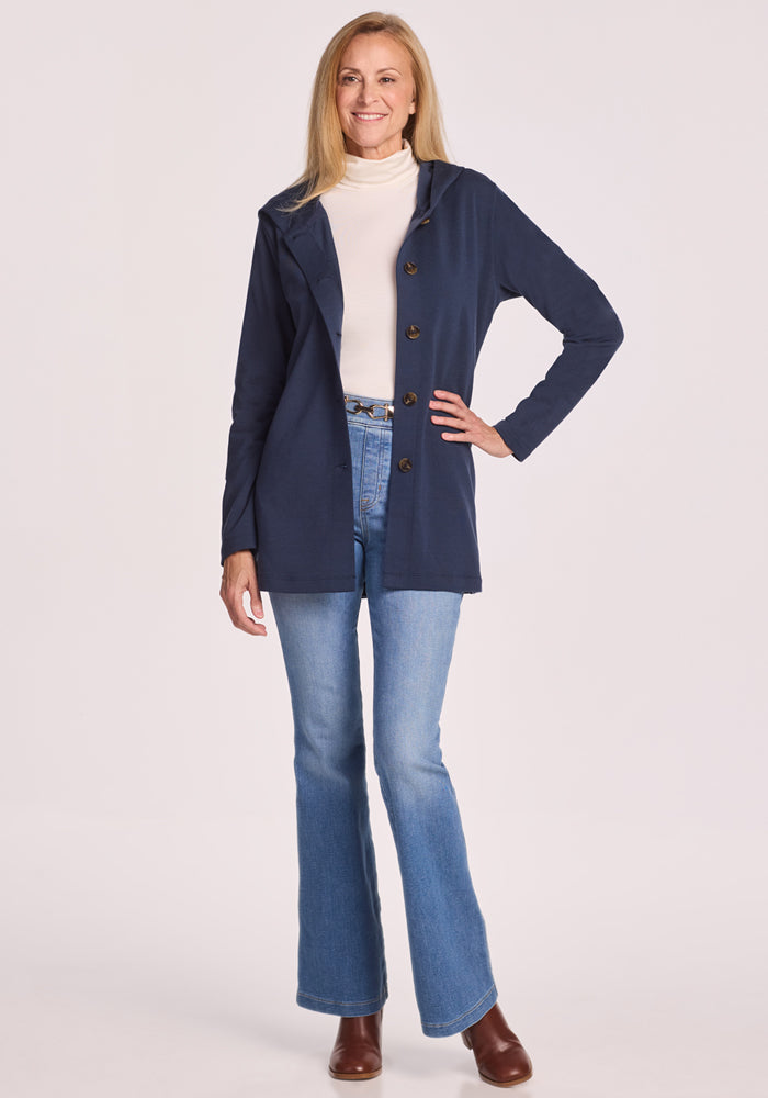 A woman with long blonde hair smiles confidently in the Woolx Edna Hooded Cardigan in Deep Navy, layered over a white turtleneck and blue flared jeans with brown boots—styled for cold days against a light background.