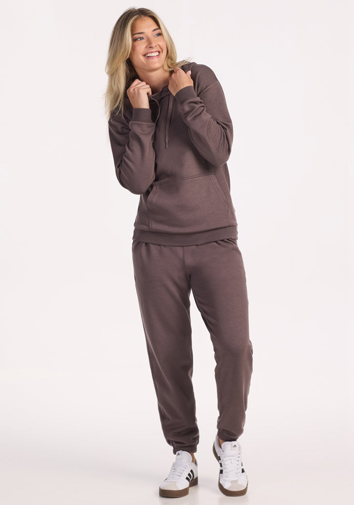 A woman smiles in a relaxed pose, wearing the Woolx Avery Hoodie in Plum Truffle with matching sweatpants and white sneakers, standing against a plain light background.