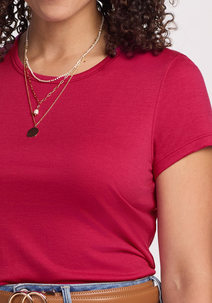 A woman wears the Woolx Addie Crew Neck in Poppy Spark, layered gold necklaces, light blue jeans, a brown belt, and has part of her curly hair visible.