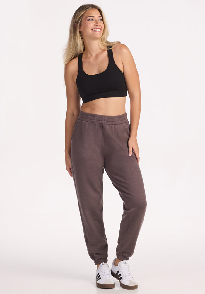 A woman with long blonde hair wears a black sports bra, Woolx Parker Sweatpants in Plum Truffle for temperature regulation, and white sneakers with black stripes, standing and smiling against a plain white background.