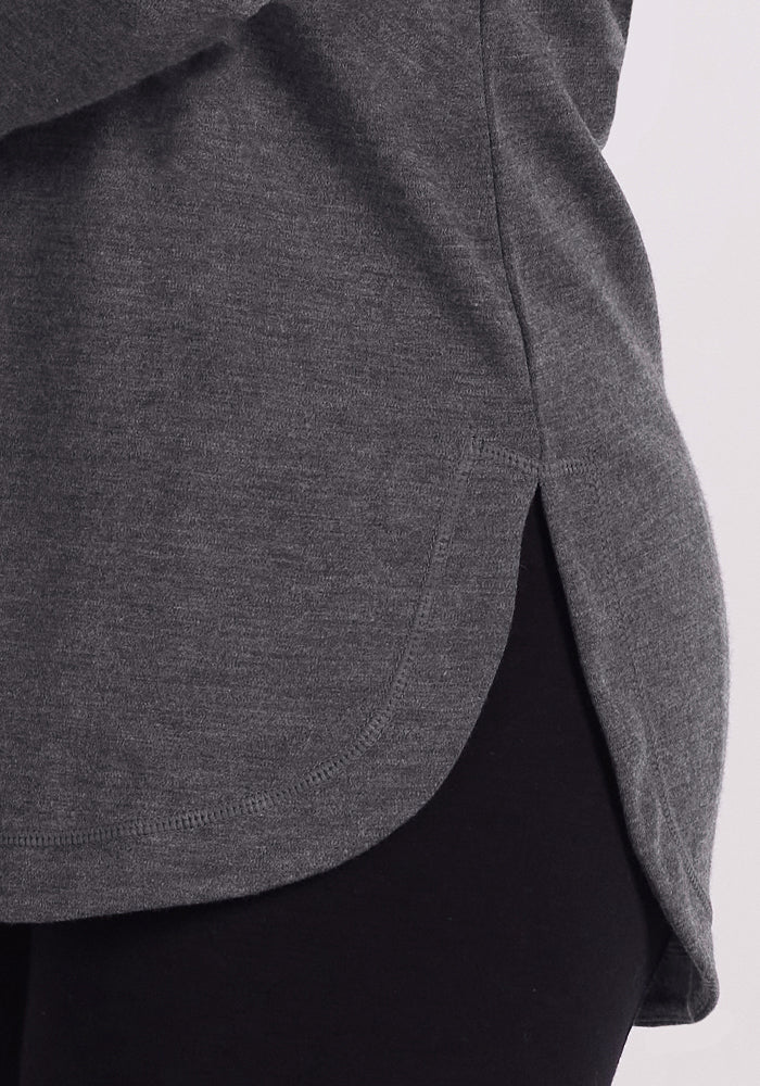Close-up of a person wearing the Woolx Blair Tunic in Cinder Melange, featuring a rounded hem and side slit. Paired with black pants, this lightweight Merino wool top provides warmth against a plain, light background.