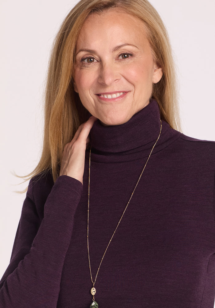 A woman with long blonde hair smiles gently at the camera, one hand touching her neck, wearing the Woolx Peyton Turtleneck in Deep Plum and a long necklace against a plain, light background.