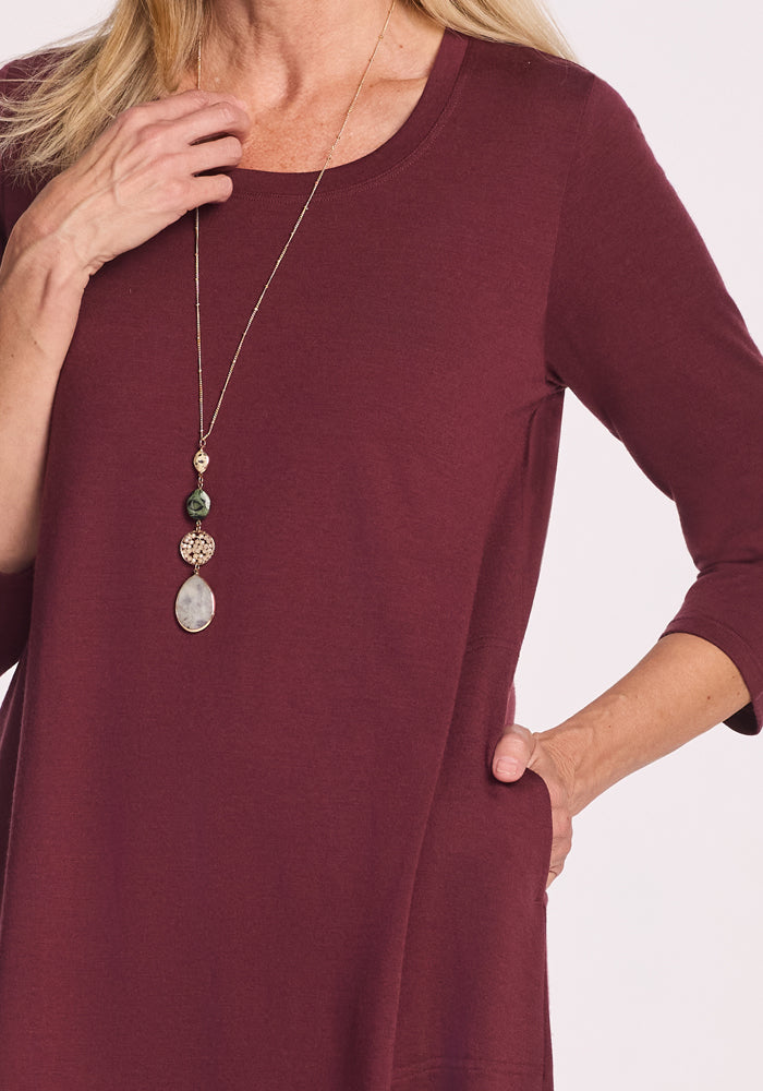 A woman models the Woolx Rissa A Line Dress in Toasted Garnet, featuring three-quarter sleeves and soft Merino wool, styled with a long beaded stone pendant necklace—perfect for fall and winter layering.