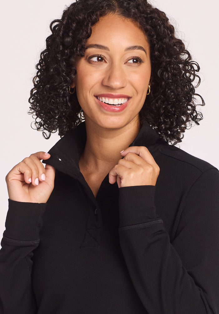A woman with curly dark hair smiles and looks to the side, lightly holding the collar of her Woolx Annie 3 Button Pullover in black. The background is plain and light-colored.