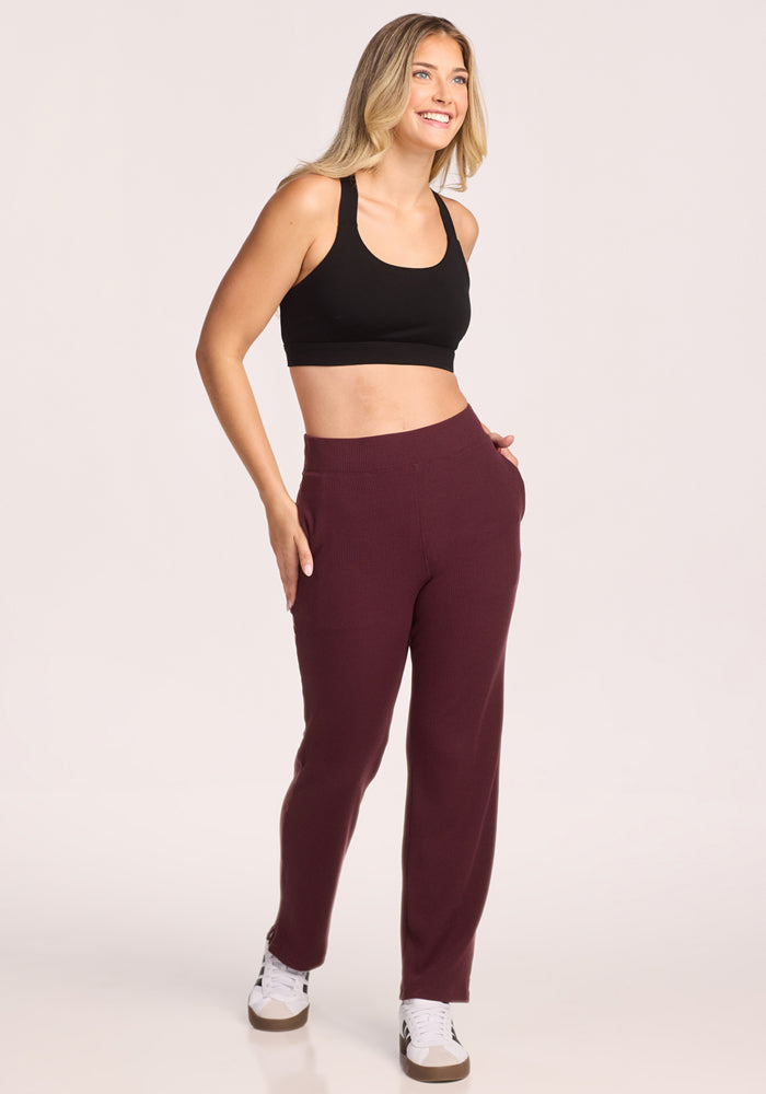 A woman with long blonde hair smiles in a black sports bra, Woolx Luca Ribbed Pants in Toasted Garnet, and white sneakers, standing with one hand in her pocket against a plain light background.