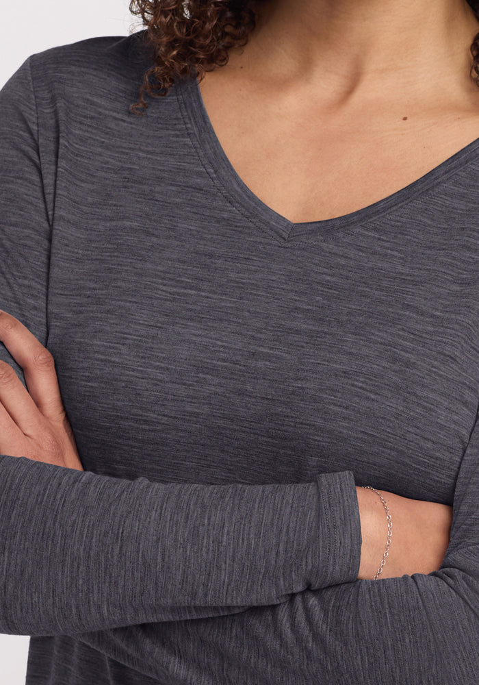A curly-haired person wears the Woolx Millie Long Sleeve in Cinder Melange, showing only their lower face, neck, chest, and arms with a thin silver bracelet. The merino wool top offers temperature regulation for comfort.