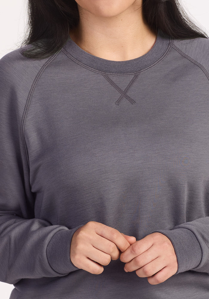 A woman wears the Woolx Bailey Crewneck Sweatshirt in Arctic Dusk, a long-sleeved gray Merino wool top, with her hands softly clasped at her waist; her face is partially out of frame and her dark hair is down.
