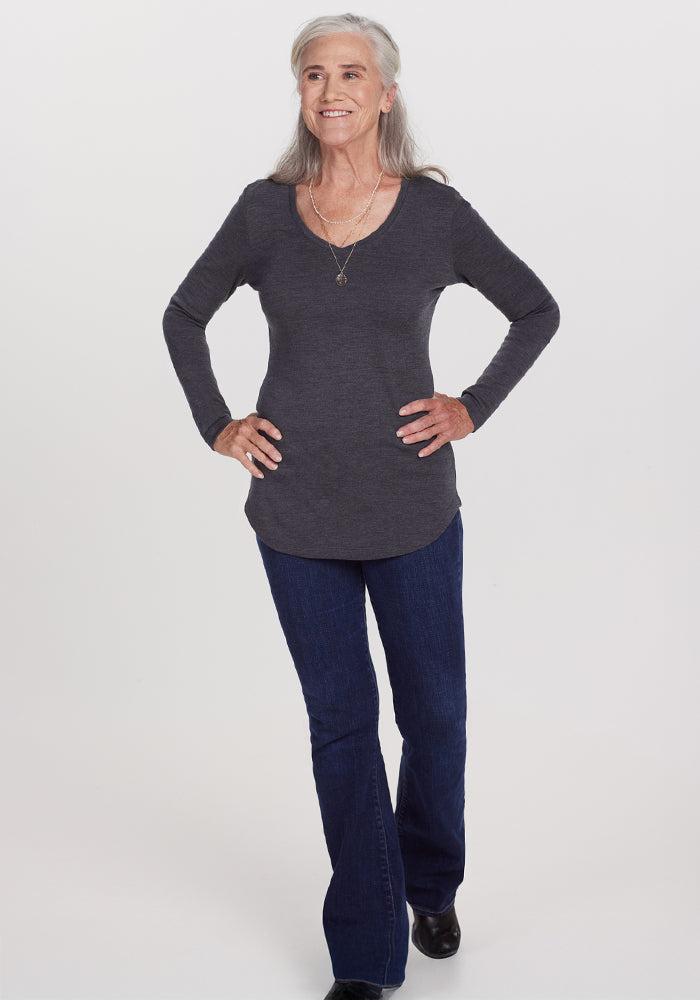 A smiling older woman with long gray hair stands confidently, hands on hips, wearing the Woolx Eva Tunic in Cinder Melange—an ideal winter essential—paired with blue jeans and a necklace, against a plain white background.