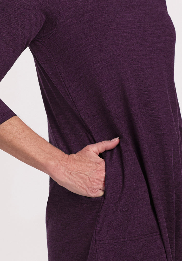 A person wearing the Woolx Rissa A Line Dress in Deep Plum, featuring three-quarter sleeves and side pockets, stands against a plain light background—an ideal Merino wool choice for stylish fall and winter layering.