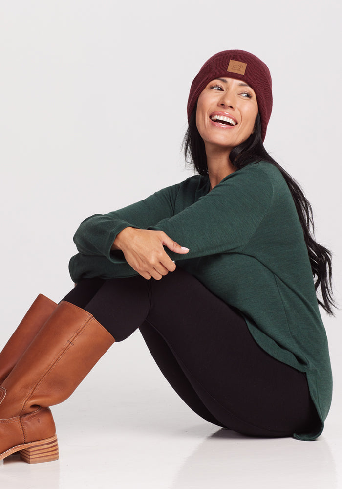 A smiling woman sits with her knees up, wearing the Woolx Blair Tunic in Forest for lightweight Merino wool warmth, paired with black leggings, tall brown boots, and a maroon beanie. Her long dark hair is down as she looks to the side.