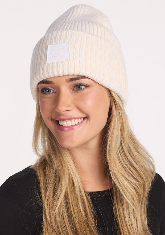 A smiling woman with long blonde hair wears the Woolx Ria Chunky Beanie in Buttercream and a black top, looking slightly to the side against a plain light background.