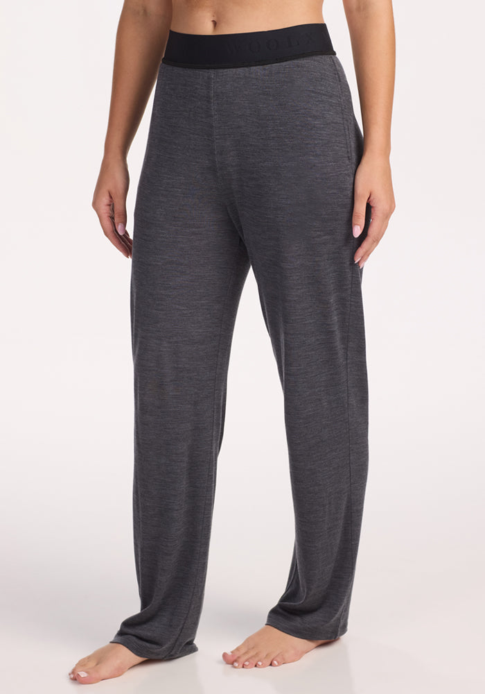 A person stands barefoot in Woolx Maya Lounge Pants in Cinder Melange, featuring a wide black waistband. The image is cropped at the shoulders against a plain, light background. 