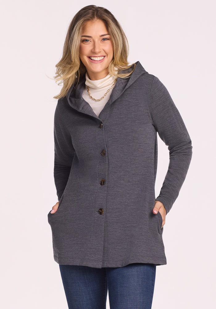 A smiling blonde woman wears the Woolx Edna Hooded Cardigan in Cinder Melange over a white turtleneck and jeans, perfect for cold weather. She stands with her hands in her pockets against a plain, light background. 