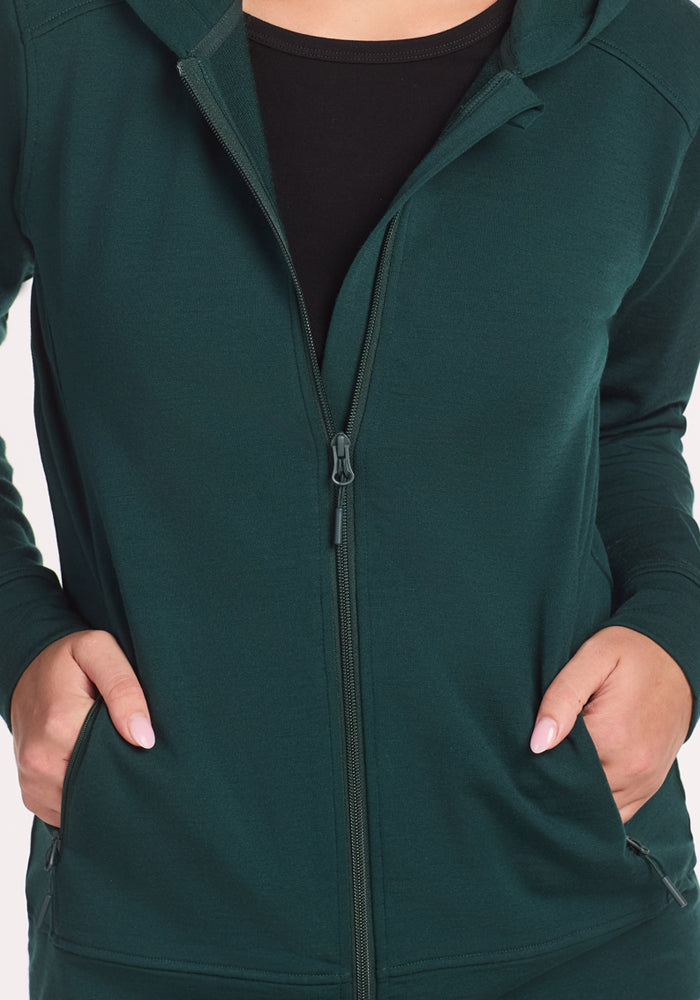 A person wearing the Woolx Darby Zip Up Hoodie in Deep Timber over a black shirt, with both hands in the hoodie pockets. Only their torso and hands are visible.