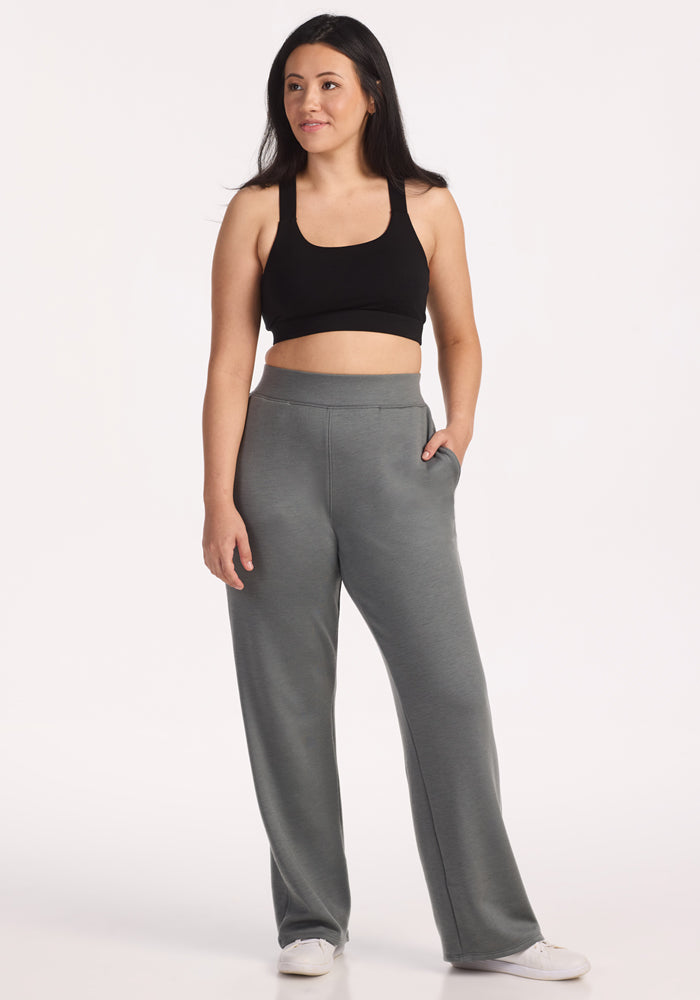 A woman with long dark hair poses against a plain background, wearing a black sports bra, Woolx Bree Lounge Pants in Sedona Sage, and white sneakers. She stands with one hand in her pocket, looking slightly to the side.