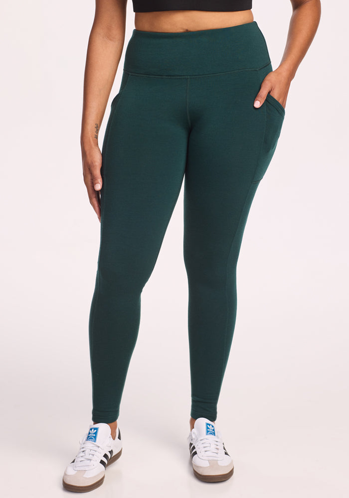 Model is wearing Woolx Piper Pocket Petite leggings in Deep Timber, paired with a black cropped top and white sneakers with blue accents, standing against a plain white background.