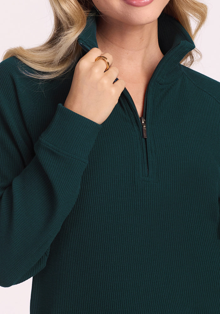 A woman with long, wavy blonde hair and a gold ring holds the collar of her Woolx Quinn Ribbed 1/4 Zip - Deep Timber top. Only her lips and chin are visible.
