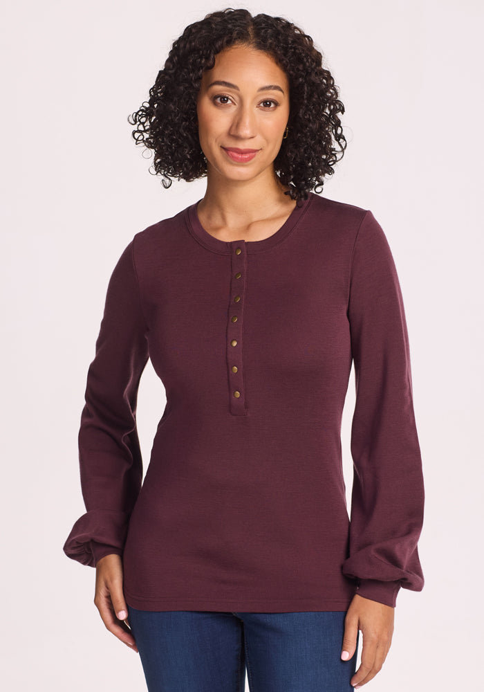 A woman with curly dark hair wears the Woolx Charlotte Puff Sleeve Top in Chocolate Fig, featuring a Henley-style button front. She stands against a plain light background, smiling gently at the camera. 