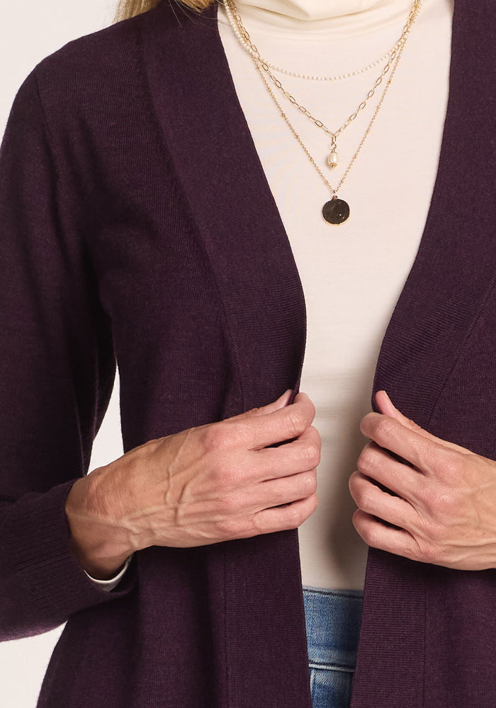 A person wearing the Woolx Macie Sweater in Deep Plum over a white turtleneck, with layered gold necklaces and blue jeans, holding the open-front cardigan at the front with both hands.