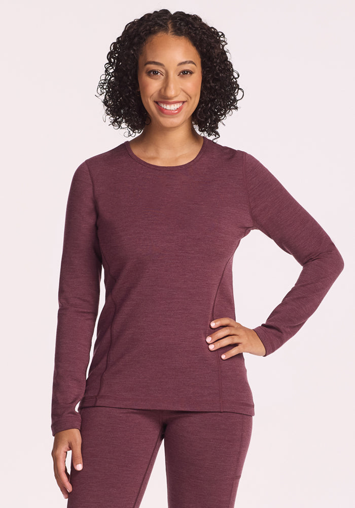 A woman with curly hair smiles, wearing the Woolx Elsa Long Sleeve in Grape Harvest Melange with matching pants, standing against a light background and posing confidently to highlight the set’s moisture-wicking comfort. 
